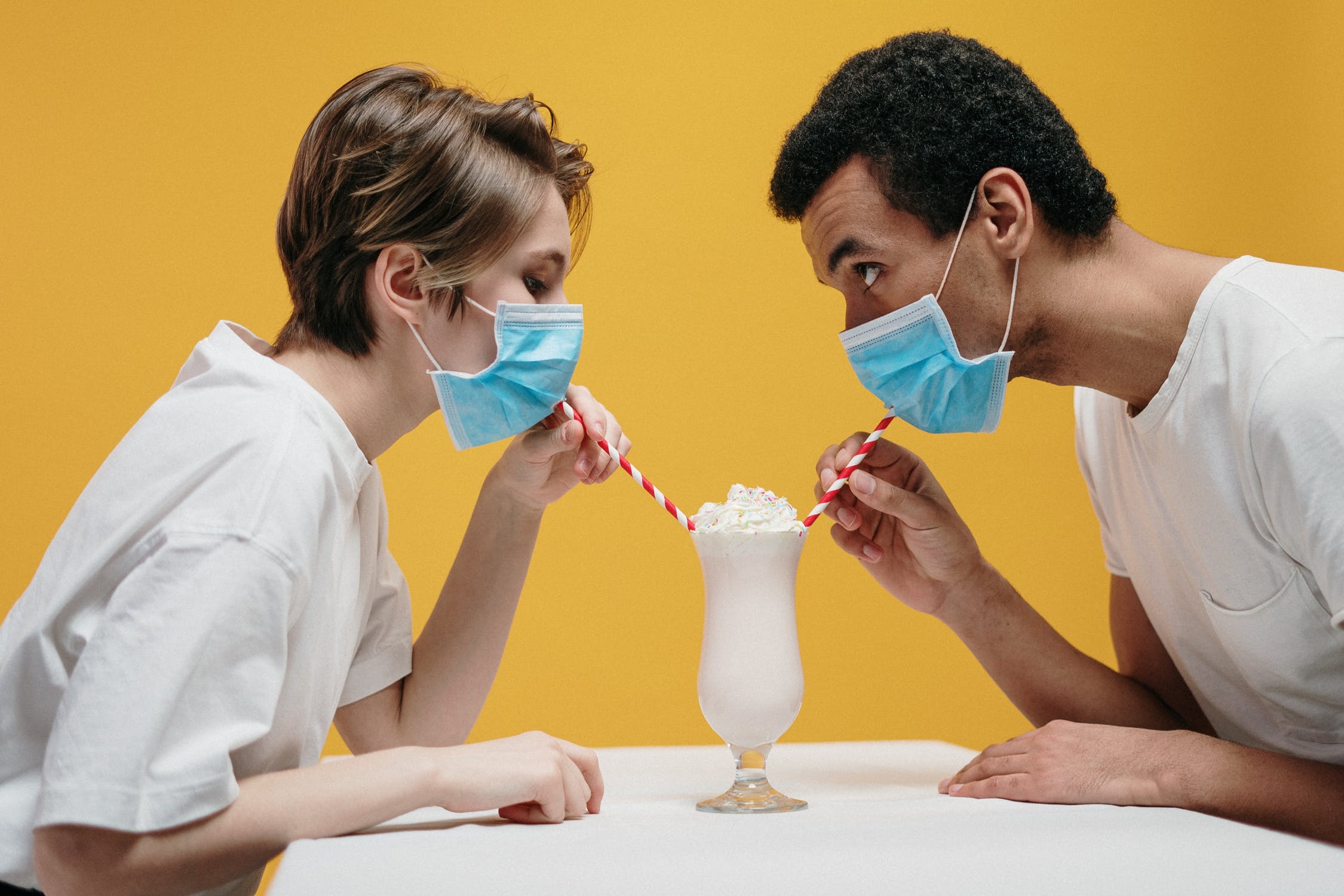 mixed race couple drinking from the same glass while wearing PPE masks against a yellow background